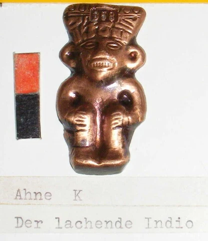 Echtes Foto: kleine Metallfigur mit Kopfschmuck und breitem Lachen, beschriftet „Ahne K – Der lachende Indio“